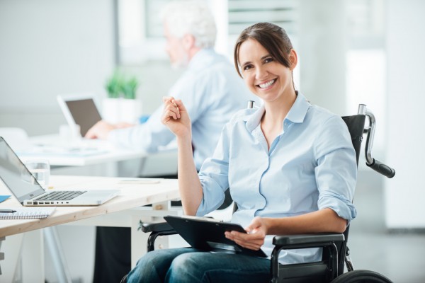 Image for inclusive recruitment: young smiling businesswoman in a wheelchair, in office environment, with laptop and tablet. 